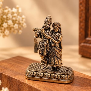 Puede incluir: Una figura de tono bronce metálico que representa a dos personas de pie juntas sobre una base rectangular con un patrón de rejilla texturizado. Una figura sostiene una flauta en sus labios, mientras la otra está a su lado, ambas con vestimenta y joyas tradicionales.