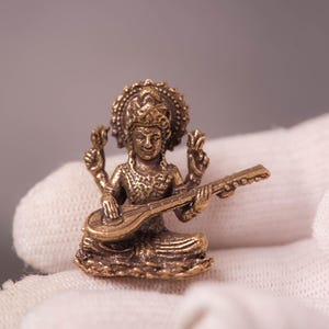 Può includere: Una piccola statua color bronzo di una figura seduta che suona uno strumento a corde. La figura ha più braccia ed è adornata da un copricapo dettagliato. La statua è tenuta in un guanto bianco, con uno sfondo neutro.