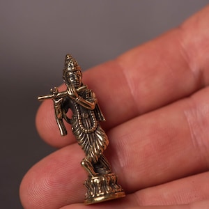 May include: A small, detailed brass figurine of a deity playing a flute. The figure is posed in a graceful stance, with intricate details in the clothing and jewelry. The figurine is held in a hand, showcasing its size and craftsmanship.