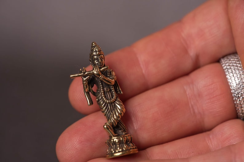 May include: A small, detailed brass figurine of a deity playing a flute. The figure is posed in a graceful stance, with intricate details in the clothing and jewelry. The figurine is held in a hand, showcasing its size and craftsmanship.