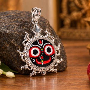 May include: A silver pendant featuring a circular enamel inlay of the Jagannath deity face with large white eyes, a red background, and a black face. The silver frame has a sunburst design with pointed rays and a decorative bail at the top.