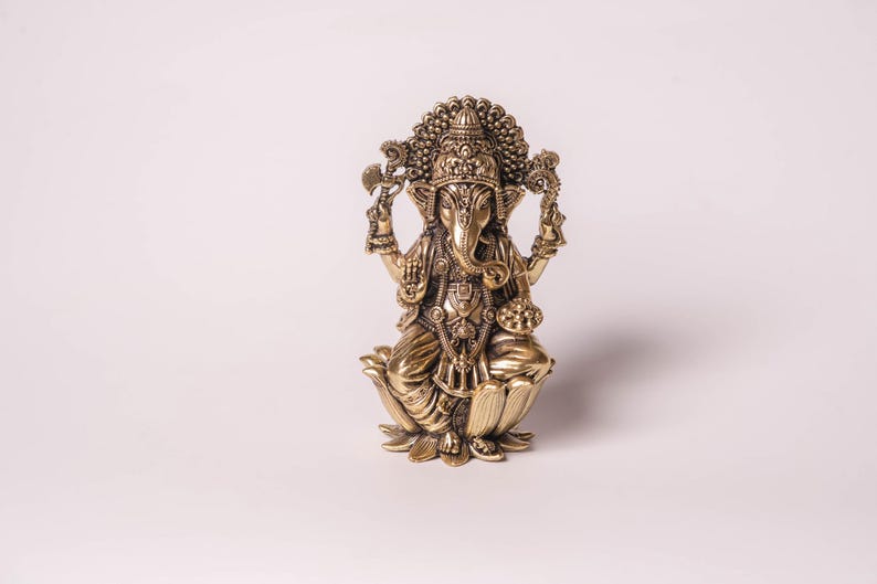 May include: A detailed golden statue of the Hindu deity Ganesha, seated on a lotus flower. The statue features intricate carvings, a large head with elephant features, and multiple arms holding various objects. The figure is adorned with ornate jewelry and a crown.