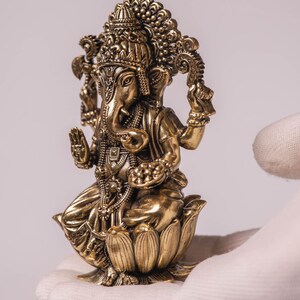May include: A detailed gold-colored statue of the Hindu deity Ganesha, seated on a lotus flower. The statue features intricate carvings, including a crown, jewelry, and multiple arms. The figure is held in a gloved hand against a white background.
