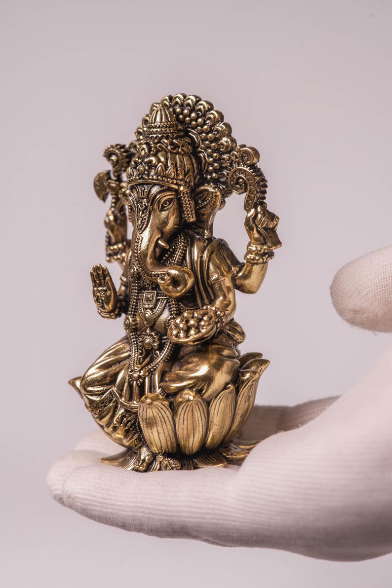 May include: A detailed gold-colored statue of the Hindu deity Ganesha, seated on a lotus flower. The statue features intricate carvings, including a crown, jewelry, and multiple arms. The figure is held in a gloved hand against a white background.