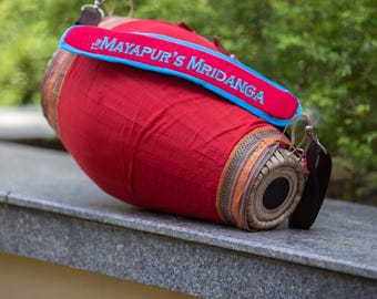 Könnte beinhalten: Eine rote Mridanga-Trommel mit einem blau-roten Riemen mit der Aufschrift "Mayapur's Mridanga" in blauer Schrift. Die Trommel hat ein dekoratives Band aus Orange, Gold und Blau an den Rändern. Die Trommel liegt auf einer grauen Oberfläche.