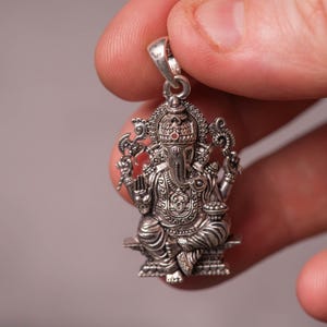 May include: A detailed silver pendant depicting the Hindu deity Ganesha, seated with multiple arms and an elephant head. The pendant is intricately designed with fine details, including a crown and ornate clothing. The pendant is held up by a silver loop.