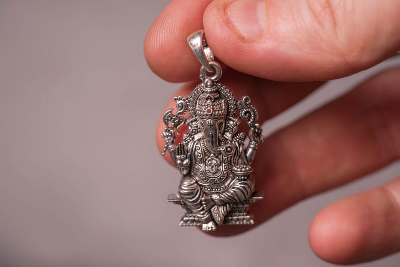 May include: A detailed silver pendant depicting the Hindu deity Ganesha, seated with multiple arms and an elephant head. The pendant is intricately designed with fine details, including a crown and ornate clothing. The pendant is held up by a silver loop.