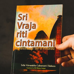 May include: A book titled "Sri Vraja Riti Cintamani" with an orange and yellow cover, featuring text and illustrations of temples and landscapes. The book is held by a hand against a dark background.