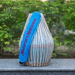 May include: A handcrafted mridanga drum with a blue strap. The drum is made of light-colored wood strips and has a blue and red interior. The blue strap is printed with the text "MAYAPUR'S MRIDANGA" in red.