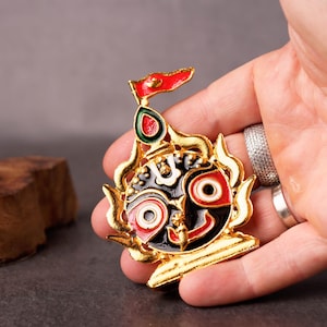 May include: A gold-colored enamel pin featuring a deity with a red flag. The pin has a black, red, and white face with a gold outline. The pin is held in a hand against a dark background.
