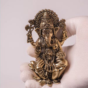 May include: A detailed brass statue of the Hindu deity Ganesha, seated on a lotus flower. The statue features intricate carvings, a large head, and multiple arms holding various objects. The figure is held in a gloved hand against a white background.