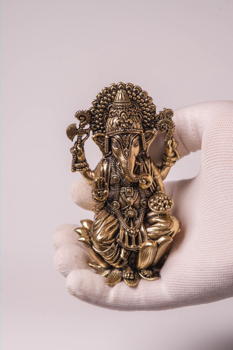 May include: A detailed brass statue of the Hindu deity Ganesha, seated on a lotus flower. The statue features intricate carvings, a large head, and multiple arms holding various objects. The figure is held in a gloved hand against a white background.