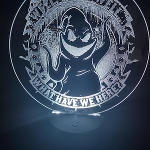 May include: A circular, illuminated acrylic night light featuring a grinning cartoon character with the text "WELL, WELL, WELL... WHAT HAVE WE HERE?" etched around the edge. The design includes bats and other spooky elements.