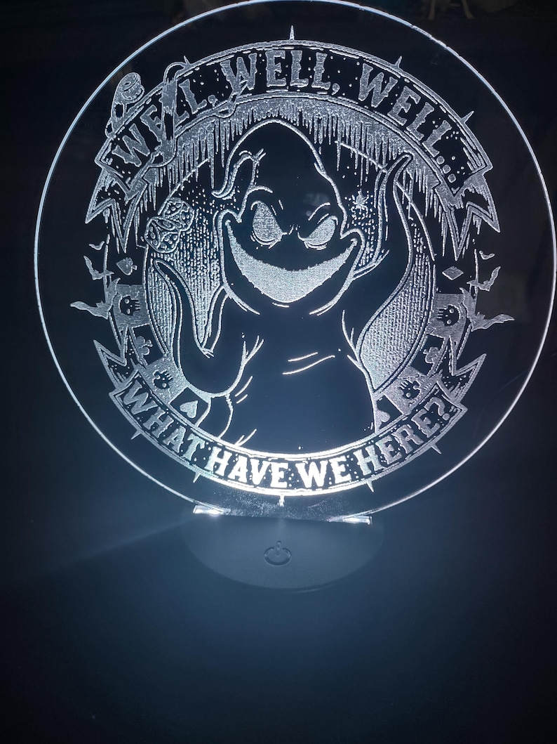 May include: A circular, illuminated acrylic night light featuring a grinning cartoon character with the text "WELL, WELL, WELL... WHAT HAVE WE HERE?" etched around the edge. The design includes bats and other spooky elements.