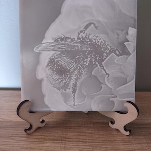 May include: A square, light-colored decorative tile featuring a detailed image of a bee on a flower. The tile is displayed on a small, wooden easel. The image is in grayscale, with the bee and flower in shades of gray and white.