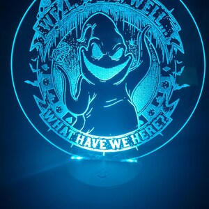 May include: A round, illuminated acrylic sign with a blue glow. The sign features a cartoon character with the text "Well, Well, Well... What Have We Here?" in a circular design. The design includes bats and other spooky elements.