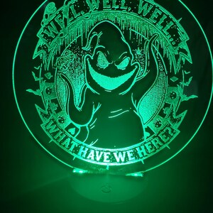 May include: A green-glowing, circular decorative light featuring the character Oogie Boogie from The Nightmare Before Christmas. The text "WE'LL, WELL, WELL, WHAT HAVE WE HERE?" is etched around the character's image.