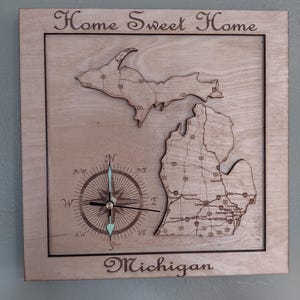 May include: Wooden wall clock featuring a map of Michigan with a compass design. The clock is engraved with the words "Home Sweet Home" and "Michigan". The clock hands are turquoise. The clock is made of light-colored wood.