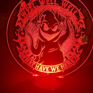 May include: A red, circular LED lamp featuring a cartoon character with the text "Well, Well, Well, What Have We Here?" The lamp has a base and emits a red glow, creating a spooky ambiance. The design is etched into the acrylic.