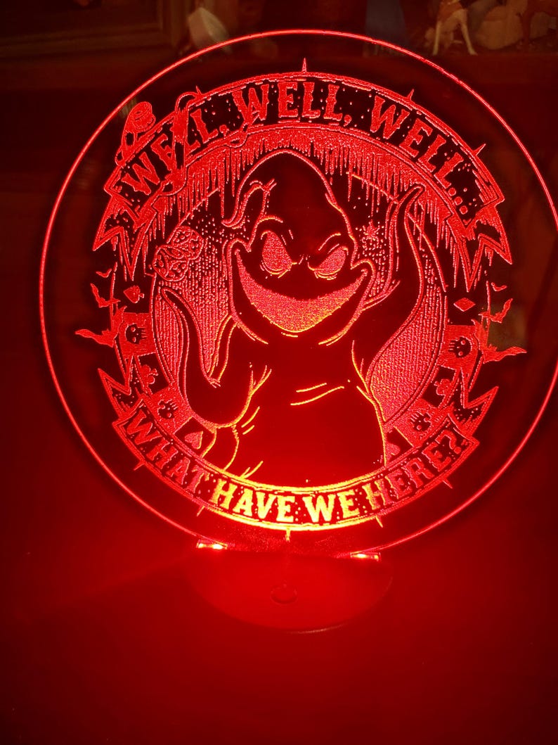 May include: A red, circular LED lamp featuring a cartoon character with the text "Well, Well, Well, What Have We Here?" The lamp has a base and emits a red glow, creating a spooky ambiance. The design is etched into the acrylic.