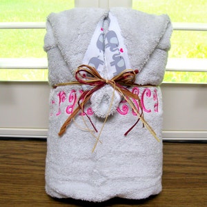 May include: A gray hooded baby towel, wrapped and tied with raffia and a decorative ribbon. The towel features pink embroidered text and a patterned band with gray elephants. The towel is folded and presented as a gift.