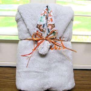 May include: A light gray hooded towel, folded and tied with raffia. The hood features a patterned fabric with foxes and bears. The towel is embroidered with orange thread. A decorative gift for a baby or child.