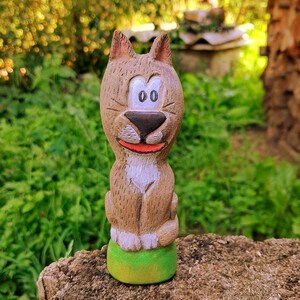 以下が含まれることがあります： 手彫りの木製猫の置物。猫は茶色の体、ピンクの耳、赤い口をしています。緑色の台座の上に座っています。置物は屋外にあり、緑に囲まれています。魅力的な装飾品です。