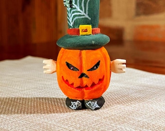 May include: A carved wooden Halloween pumpkin figure with a green top hat featuring a white spiderweb design and a small black spider. The pumpkin has an orange body, black eyes, and a carved mouth. The figure has small wooden arms and black shoes.