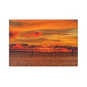 Puede incluir: Impresión rectangular en lienzo que muestra una puesta de sol sobre el océano. El cielo es una mezcla de tonos naranja, rojo y amarillo, con nubes dispersas. Dos postes oscuros se alzan en el agua, acentuando la profundidad del paisaje marino.