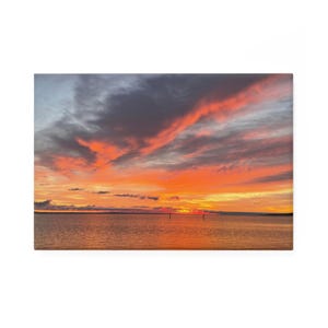 May include: A vibrant sunset over calm water. The sky is filled with streaks of orange, red, and gray clouds. The water reflects the colors of the sky, creating a serene and colorful landscape.