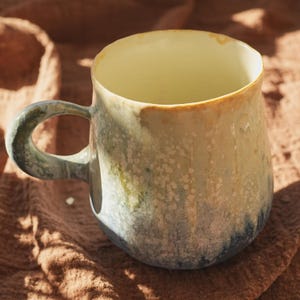 Puede incluir: Una taza de cerámica hecha a mano con un cuerpo redondeado y un asa grande. La taza presenta un esmalte moteado en tonos crema, azul, verde y marrón. El interior es de color amarillo pálido. La taza mide unos 10 cm de alto.