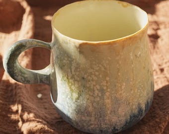 May include: A handmade ceramic mug with a rounded body and a large handle. The mug features a speckled glaze in shades of cream, blue, green, and brown. The interior is a pale yellow. The mug is approximately 4 inches tall.