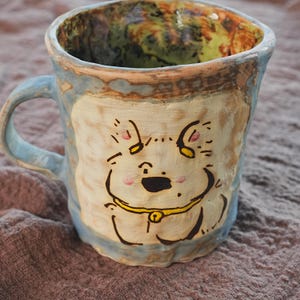May include: Hand-painted ceramic mug with a whimsical dog face design. The mug features a light blue handle and rim, with a cream-colored body. The dog's face is outlined in black, with pink cheeks and a yellow collar. The interior has a mottled green and brown glaze.
