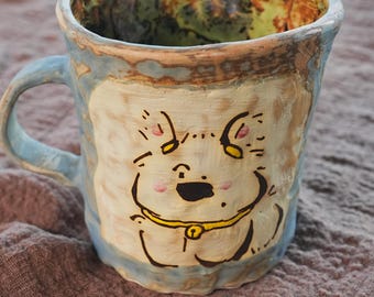 Puede incluir: Taza de cerámica pintada a mano con un diseño caprichoso de cara de perro. La taza tiene un asa y un borde azul claro, con un cuerpo de color crema. La cara del perro está delineada en negro, con mejillas rosas y un collar amarillo.