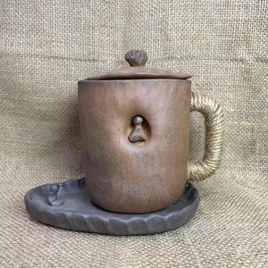 Può includere: Una tazza in ceramica marrone fatta a mano con coperchio e manico in corda. La tazza presenta un design intagliato unico e poggia su un piattino abbinato. La tazza è alta circa 10 cm.