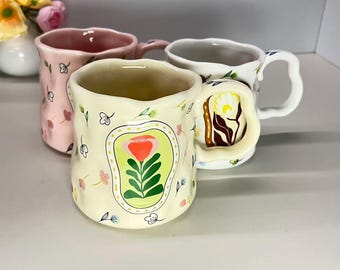 May include: Four ceramic mugs with floral designs. One mug is light yellow with a red flower and green leaves. Another is pink with small flowers. The third is white with green leaves. The fourth mug has a unique handle.