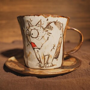 May include: A handmade ceramic mug and saucer set. The mug features a whimsical cat illustration with large eyes and a red tongue. The mug and saucer have a rustic, textured appearance with brown accents.