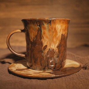 May include: A handcrafted ceramic mug and saucer. The mug features a textured, organic form with a brown and tan marbled glaze. The matching saucer is round and complements the mug's earthy tones. Perfect for enjoying tea or coffee.