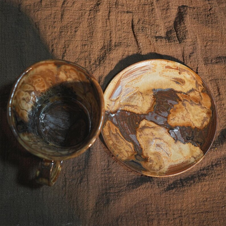 May include: Overhead shot of a handmade ceramic mug and saucer set. The mug has a handle and a dark brown interior, with a mottled brown and tan exterior. The round saucer features similar brown and tan patterns.