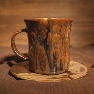 May include: A handmade ceramic mug with a distinctive glaze in brown, blue, and cream hues. The mug features a curved handle and rests on a matching saucer. The rustic design is suitable for tea or coffee.