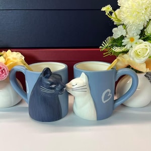 May include: Two light blue ceramic mugs with cat designs. One mug features a dark grey cat, the other a white cat, touching noses. Each mug has a gold spoon inside and a white heart design.