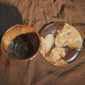 May include: A handmade ceramic mug and saucer set. The mug has a brown and tan marbled design with a handle. The saucer has a similar design. The set is on a brown fabric background. The mug is approximately 10 cm tall.