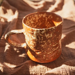 Könnte beinhalten: Eine handgefertigte Keramiktasse mit rustikaler, strukturierter Optik. Die Tasse hat eine warme, erdige Farbpalette aus Braun und Creme mit einem stabilen Henkel. Das einzigartige Design deutet auf einen handgefertigten Artikel hin.