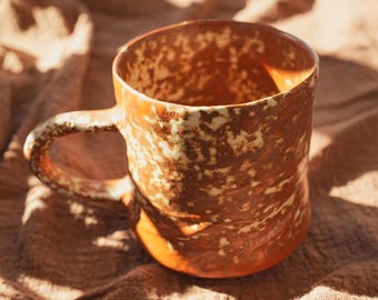 May include: A handcrafted ceramic mug with a rustic, textured finish. The mug has a warm, earthy colour scheme of brown and cream, with a sturdy handle. The unique design suggests a handcrafted item, ideal for enjoying coffee or tea.