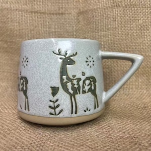 Puede incluir: Una taza de cerámica con un acabado moteado gris claro. La taza presenta un diseño de ciervo en color gris oscuro, junto con detalles florales y de copo de nieve. El asa tiene forma triangular. La base de la taza es de color beige.