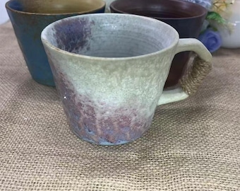 Puede incluir: Una colección de tazas de cerámica hechas a mano en varios colores. La taza delantera tiene un esmalte texturizado blanquecino con detalles morados y marrones. El asa está envuelta con cuerda natural. Otras tazas son azules, marrones y beige.