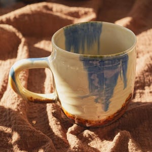 May include: A handmade ceramic mug with a cream-colored base and a blue, dripped glaze design. The mug features a rounded handle and a brown and orange rim. The mug is sitting on a textured, brown surface.