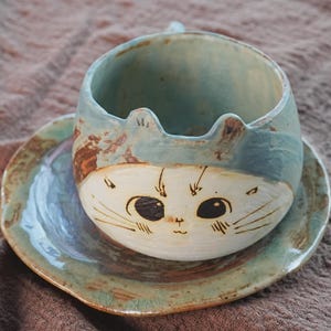 May include: A ceramic cat-shaped teacup and saucer set. The cup is light blue with a white face, black eyes, and whiskers. The saucer and cup have a rustic, handmade appearance, with brown and green accents.
