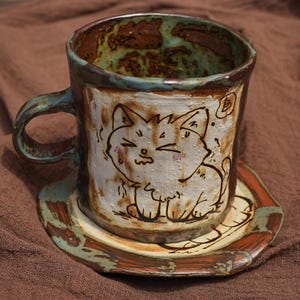 Puede incluir: Juego de taza y platillo de cerámica hechos a mano. La taza presenta una ilustración de gato dibujada a mano con mejillas rosadas. La taza y el platillo tienen un aspecto rústico y texturizado con acentos marrones, blancos y teal.