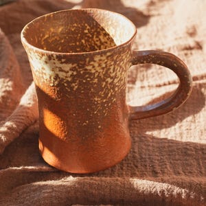 May include: A handmade ceramic mug with a rustic, speckled design. The mug features a brown and tan color scheme with a rounded handle. The mug is designed for hot beverages and is suitable for home use.